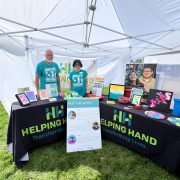 Two people stand behind tables under a white canopy. The tables display artwork, photos, and flyers. Signs read "Helping Hand: Transforming Lives." The setting is outdoors on a grassy area.