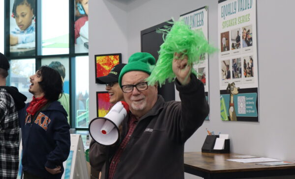 An older man wearing a green hat and glasses smiles while holding a green pom-pom and a megaphone indoors. Other people are standing nearby, and colorful posters are visible on the walls behind them.