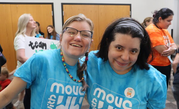 Two women smiling and posing together in matching blue "Move with Us" shirts at an indoor event, surrounded by other people. One woman wears glasses and a colorful bead necklace.