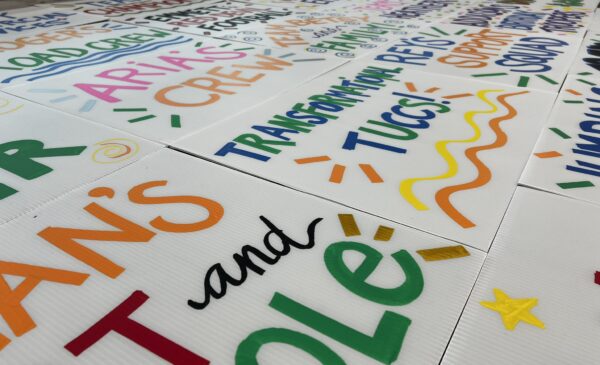 Colorful hand-painted signs with bold letters and decorative elements like stars and swirls are spread out on a flat surface, featuring phrases such as "TRANSFORMATION," "TUCCI," and "CREW.