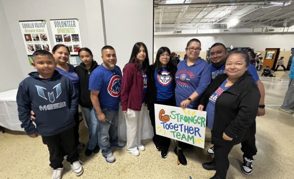 A group of people stand indoors, smiling and holding a colorful sign that reads "Stronger Together Team." They are dressed casually and appear to be at a community event.