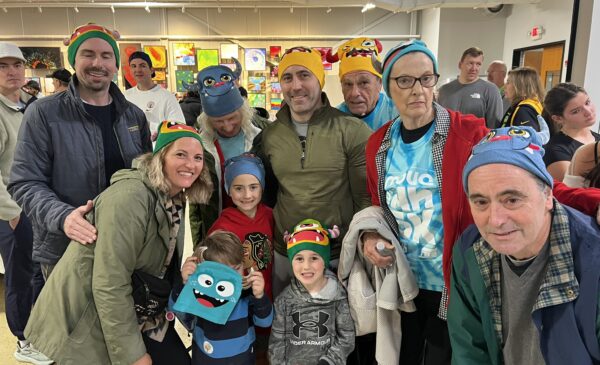A group of people, including adults and children, pose together indoors wearing colorful character hats. They are smiling and appear to be at an event or gathering, with art displayed on the walls behind them.