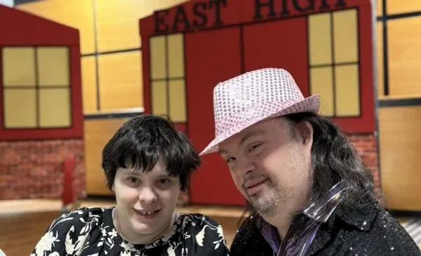 Two people smile together indoors in front of a red and yellow "EAST HIGH" stage backdrop. One wears a black-and-white floral top, the other a sparkly jacket and a pink hat.