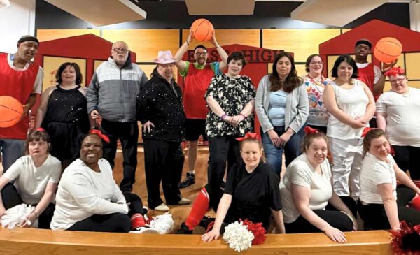 A group of people pose indoors, some dressed as basketball players with orange basketballs, others as cheerleaders holding pom-poms. They are smiling and standing on a wooden floor with a school gym backdrop.