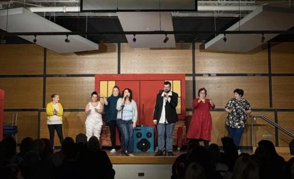 Seven people stand on a stage performing in front of an audience; they hold microphones and wear varied casual and semi-formal outfits, with a wooden backdrop and stage lighting overhead.
