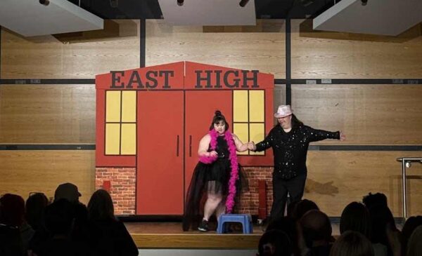 Two performers are on stage in front of a backdrop reading "EAST HIGH." One wears a black dress and pink feather boa, the other a sparkly black outfit and white hat. An audience watches them from the darkened auditorium.