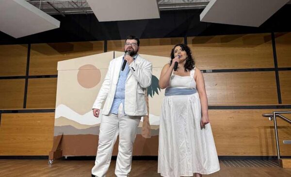 Two people stand on a wooden stage, holding microphones and singing. Both are dressed in white; the man wears a white suit with black shoes, and the woman wears a white dress and sandals. A painted backdrop is behind them.