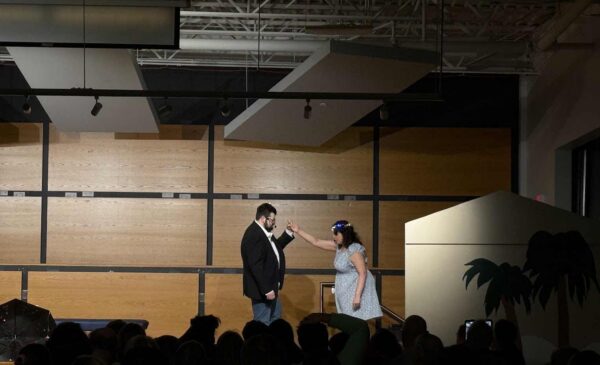 A man in a suit and a woman in a white dress stand facing each other on a stage, with the woman holding a microphone towards the man. An audience watches in a dimly lit room with a wood-paneled backdrop.