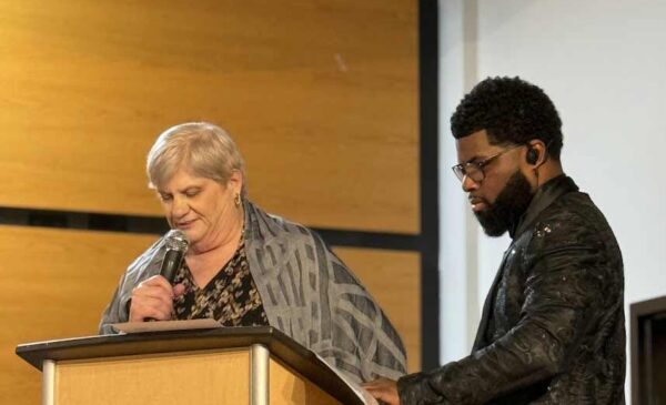 A woman and a man stand at a podium indoors. The woman holds a microphone and reads from a paper, while the man stands beside her looking at the podium. Both are dressed formally.