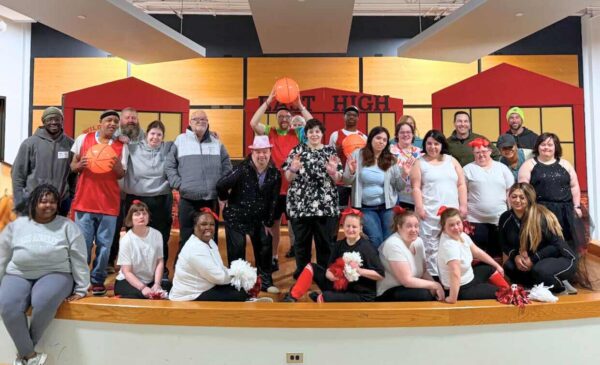 A diverse group of adults and young people pose together on a stage, smiling. Some hold basketballs or pom-poms, and several wear matching shirts. The background features a sign that says “HIGH.”.