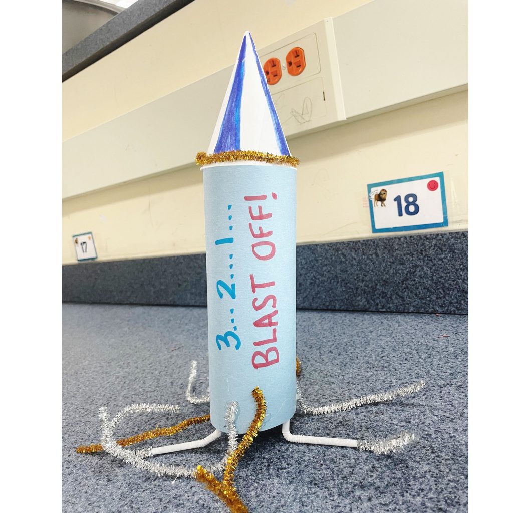 This image shows a bottle rocket created from a water bottle, bamboo sticks, pipe cleaners, and construction paper. The side of the bottle rocket says, "3...2...1...Blast Off!"