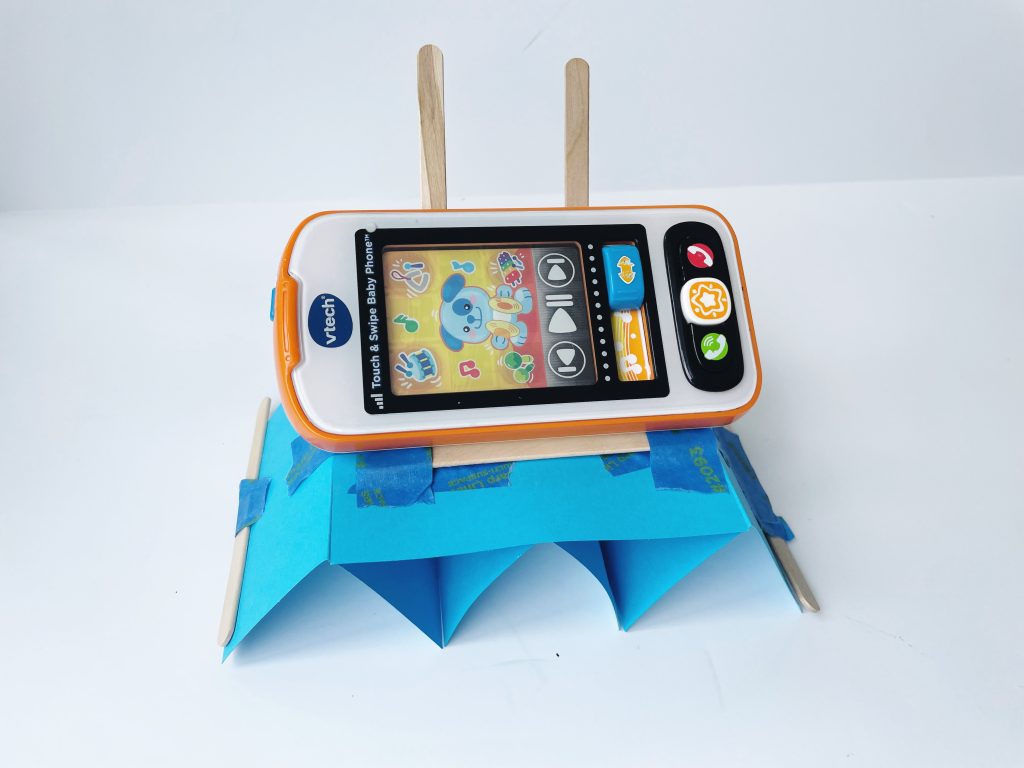 This image shows a toy phone sitting on a DIY cell phone stand created from wooden craft sticks, pipe cleaners, cardstock, and tape.