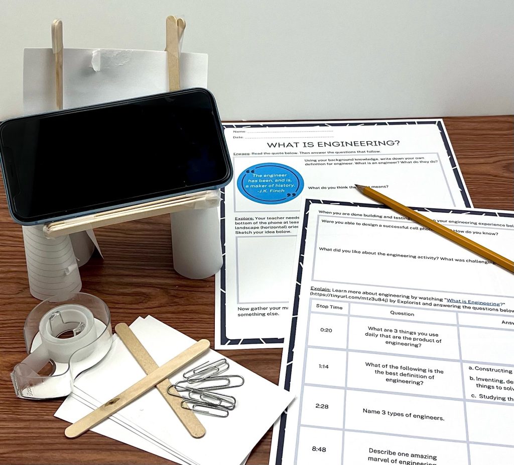what is engineering lesson materials worksheets, pencil, tape, popsicle sticks, flash cards, and paper clips and phone on a designed phone stand with dark wood desk background