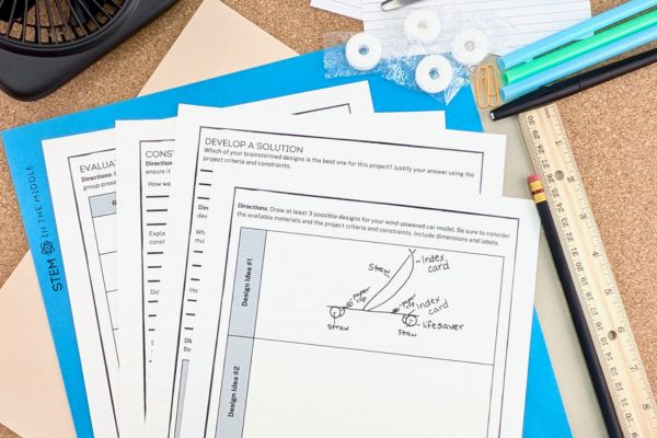 This image shows a sketch for a wind-powered car design. The worksheets are surrounded by a fan and household supplies used to build the car.