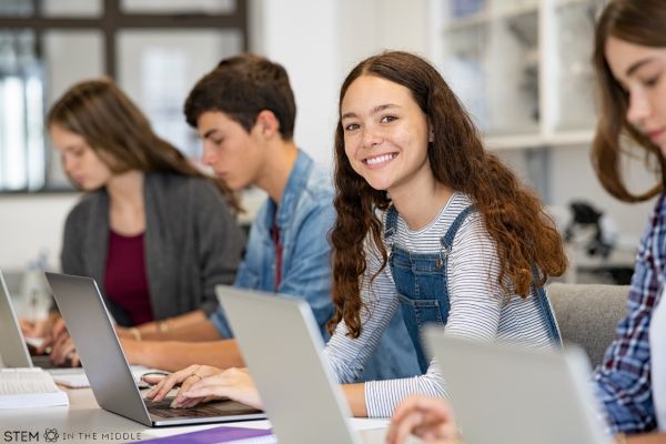 This image shows four teenagers with laptops in a classroom setting. Three students are looking at their devices. One is smiling at the camera.
