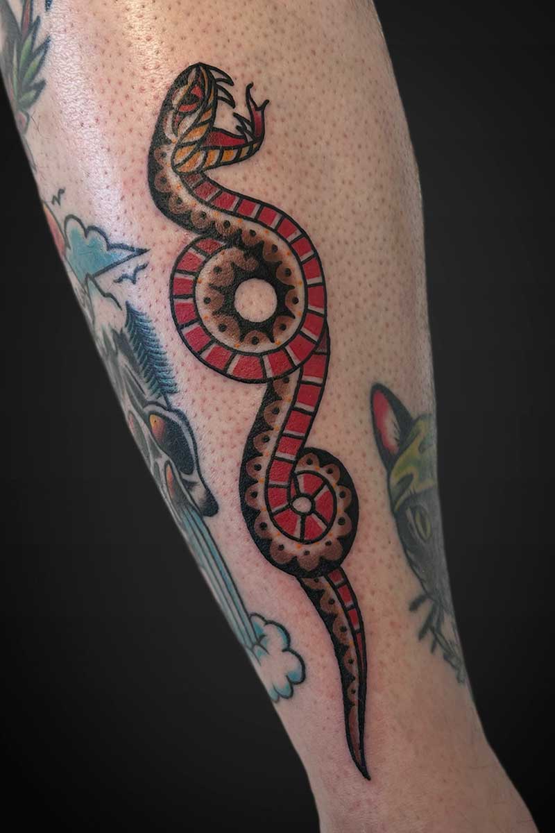 Colorful tattoo of a stylized snake winding down a forearm, with red segments and black outline, brown shading, and a curled tail near the wrist.