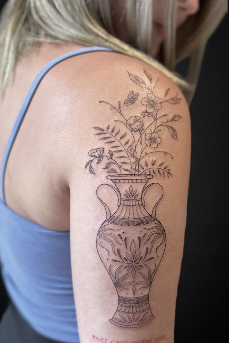 Line-drawn vase with blooming flowers tattoo on the upper arm/shoulder, in black ink.