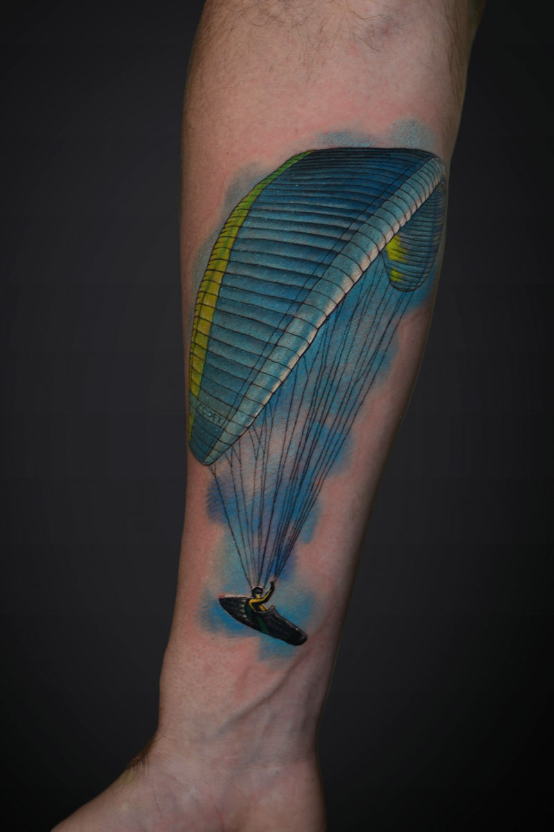 Colorful tattoo of a person paragliding under a blue-yellow canopy along the forearm.