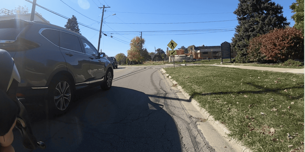 An SUV rolls just on the left as we look down a street with lawn, bushes and trees on the right and a pedestrian crosswalk ahead.