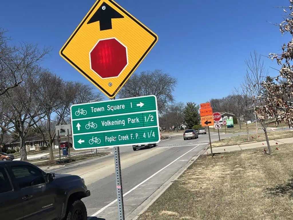 A yellow diamond sign indicating a stop sign coming up, with three horizontal bike route labels, is seen before a bike lane on a paved neighborhood road shortly before an intersection with construction signage on the right.