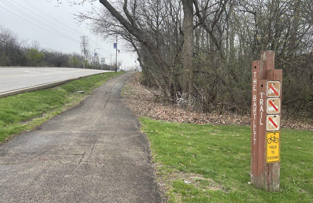 Looking down a rough paved trail, off the side of a fairly open road, with leafless trees and bushes in the background, and a wood post sign that says The Bartlett Trail.