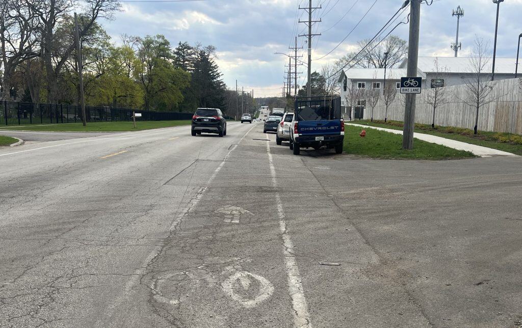 Looking down a bike lane on a street with cracks and debris, through an industrial area with cars and trucks parked on the right, some trees in the left background.