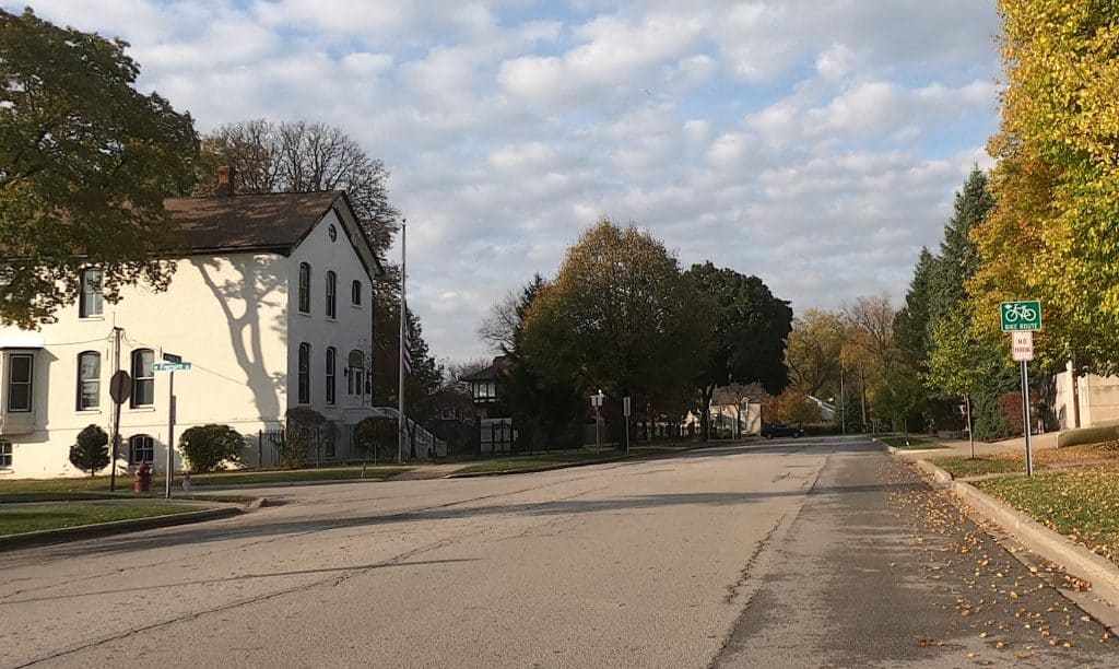 Looking down a wide street through a suburban neighborhood with a big white house on the left just beyond an intersection and trees in the background, and a Bike Route sign on the right with an arrow pointing left.