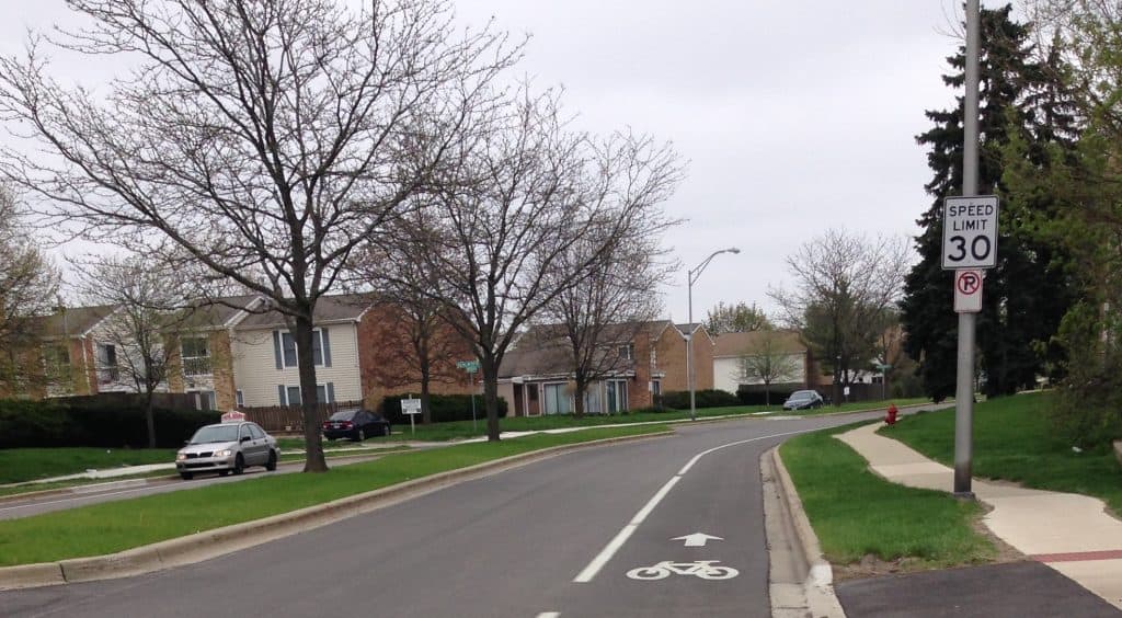 Looking down a newly paved black road with a bike lane marked with a bicycle drawing and an arrow, through a suburban neighborhood of townhomes, leafless short trees around.