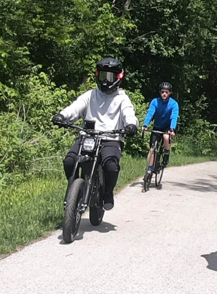 An e-bike rider with a motorcycle-like helmet rides toward us with a regular bicyclist behind on a paved forest bike trail.