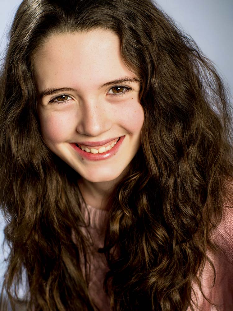 Natural smile headshot of young woman with long, curly brown hair against soft grey background, professional Sussex headshot photography for corporate or personal branding.