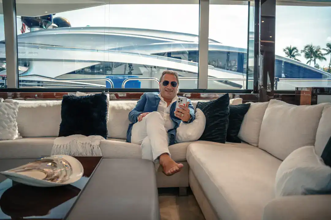 Luxurious man relaxing on a white sofa inside a yacht with large windows showcasing yacht marina views, emphasizing premium yachting experiences by Syslo Ventures.