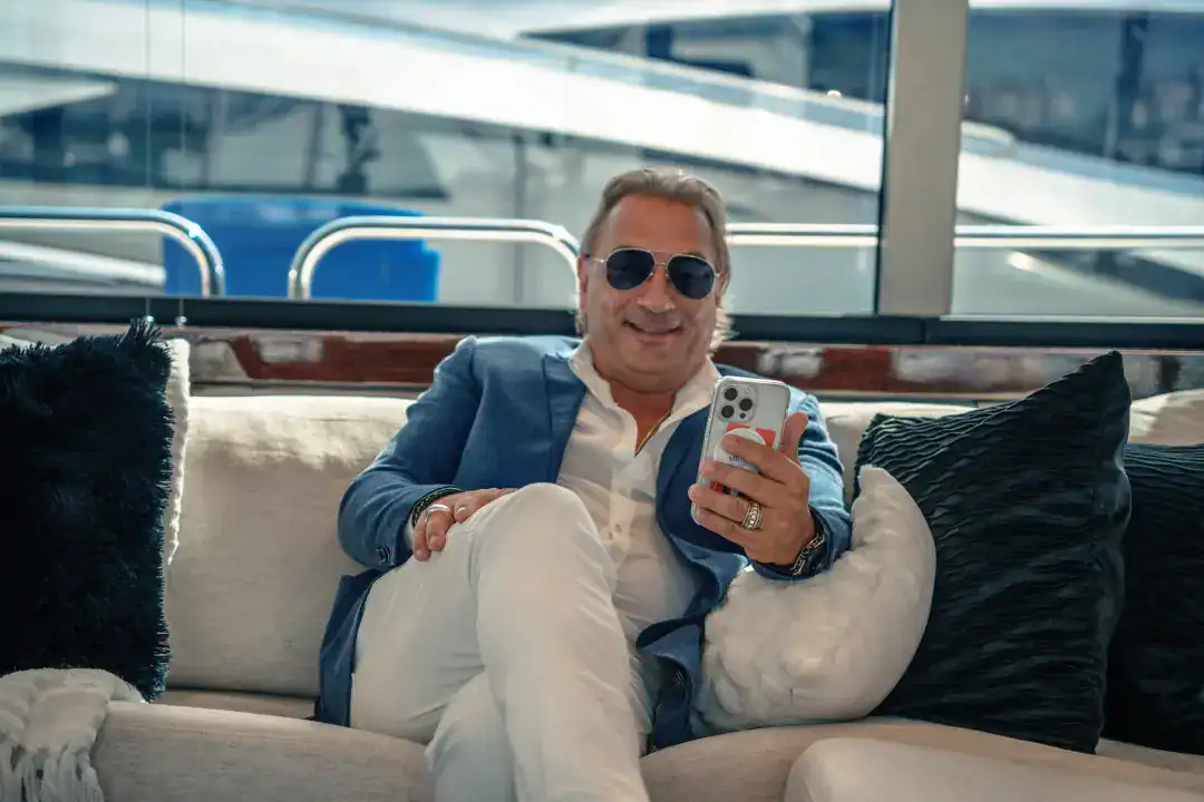 Relaxed man in sunglasses enjoying mobile phone on luxury yacht at marina, representing upscale lifestyle and marine travel.