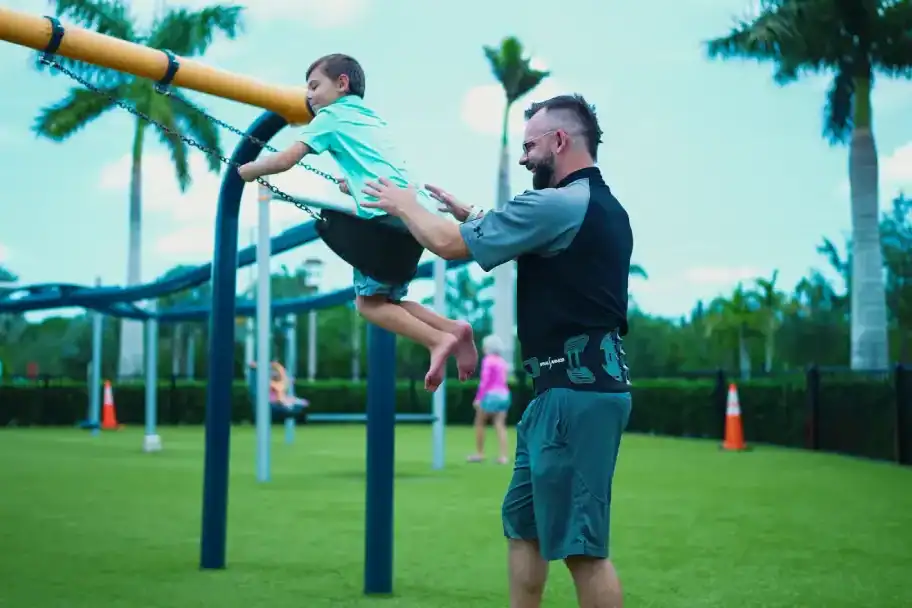 Child playing on outdoor fitness equipment at park, supervised by adult man, family outdoor activity, healthy lifestyle, community recreation, tropical park setting, playground, kids exercise, fun outdoor fitness, summer day.