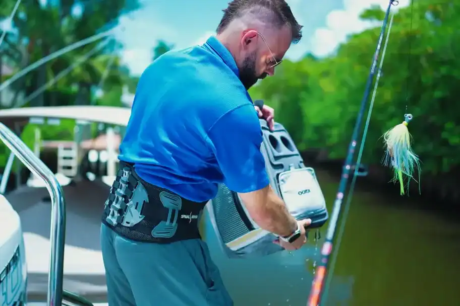 2. Man in blue fishing shirt on boat holding tackle box by water with fishing rod and lure in background.