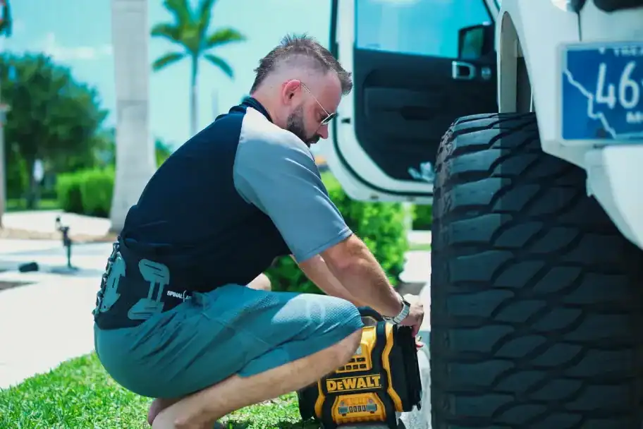 DeWalt cordless power drill being used by a man fixing a large off-road vehicle outdoors, with palm trees and bright sky in the background.