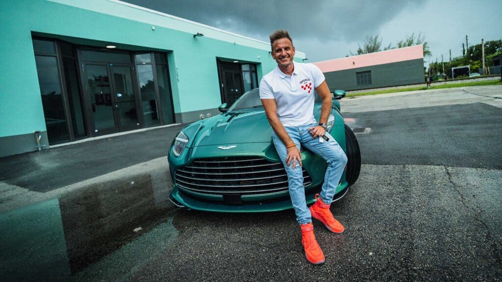 Luxury sports car parked outside a modern building with a smiling man sitting on its front, showcasing entrepreneurship, success, and high-end investments.