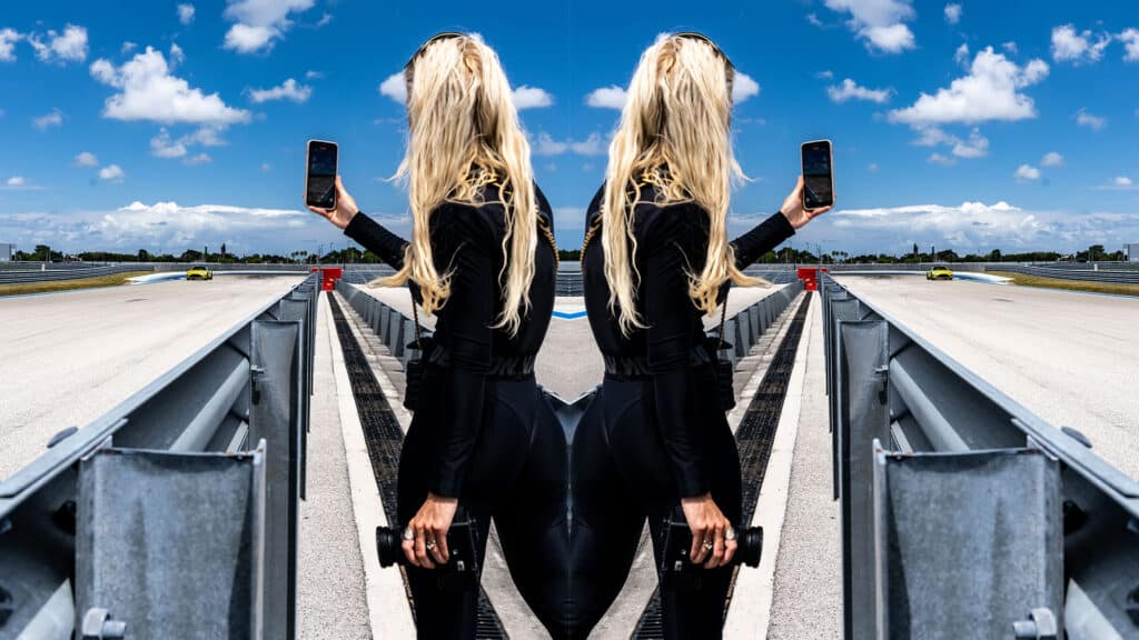 Female photographer at race track holding smartphone and camera, with edited mirrored effect, sunny day, blue sky with clouds, racing cars in background, professional motorsport photography, high-speed racing environment, dynamic action shot, motorsport event, trackside photography.