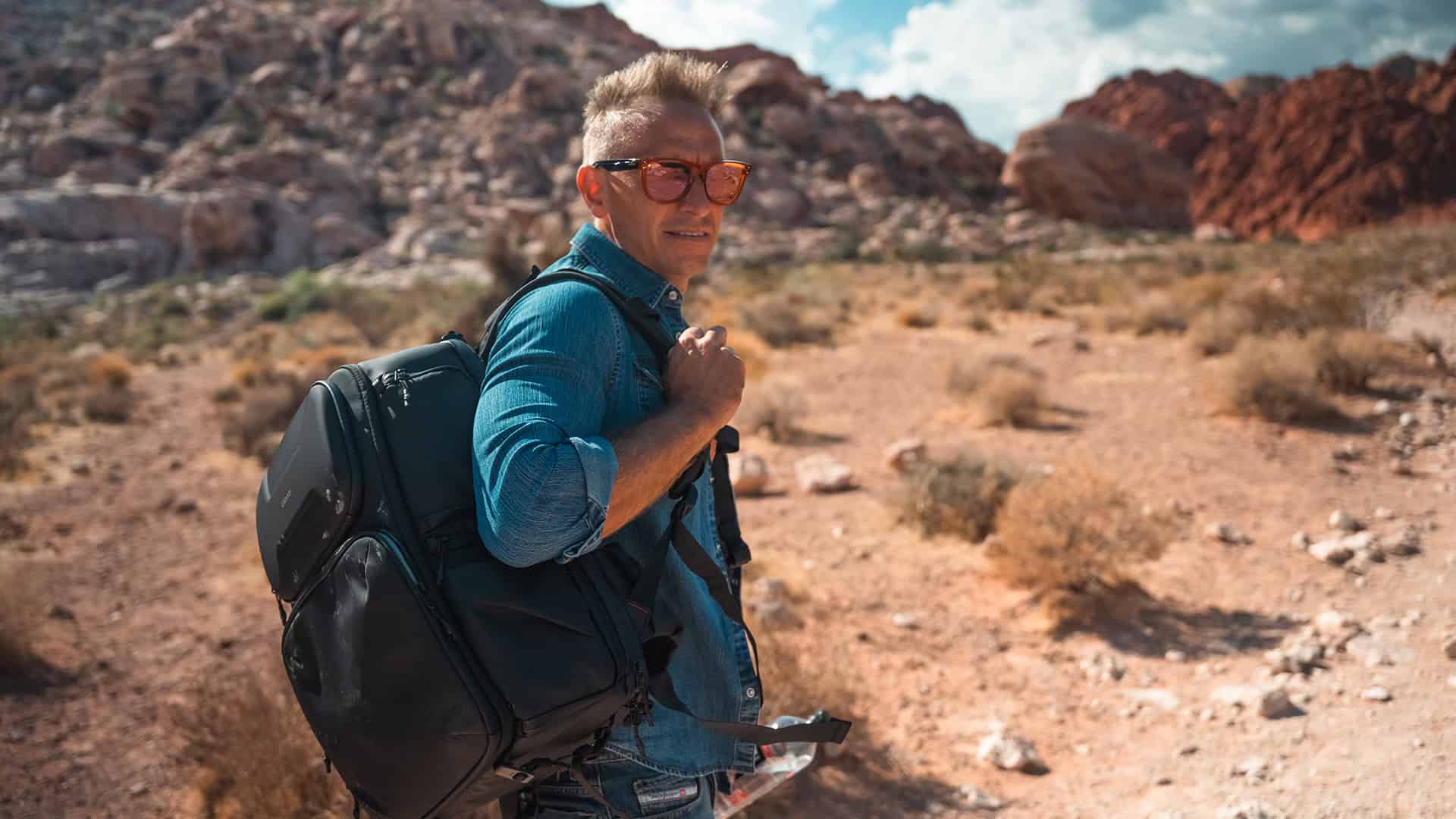 Traveling man with backpack hiking through desert landscape with red rocks and blue sky, wearing sunglasses, exploring outdoor adventure, representing outdoor gear and adventure travel.