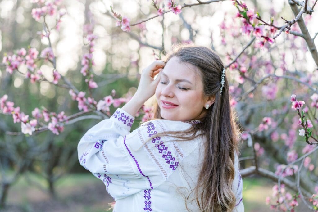 Expecting mother closes her eyes and smiles in her traditional Ukrainian maternity dress among the peach blossoms in Charlotte NC