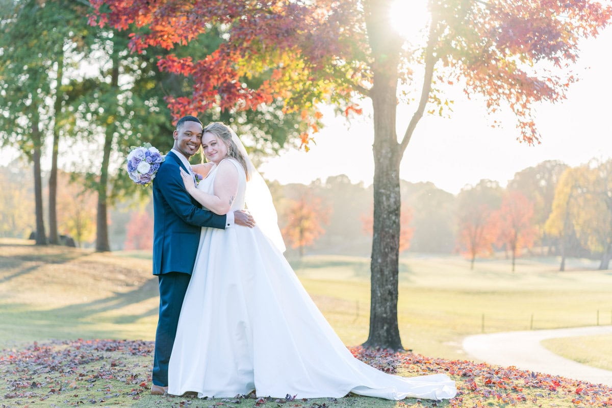 Megan and Kaylin stand under a gorgeous red tree during golden hour. The sun peaks through the trees, casting a beautiful light on them.