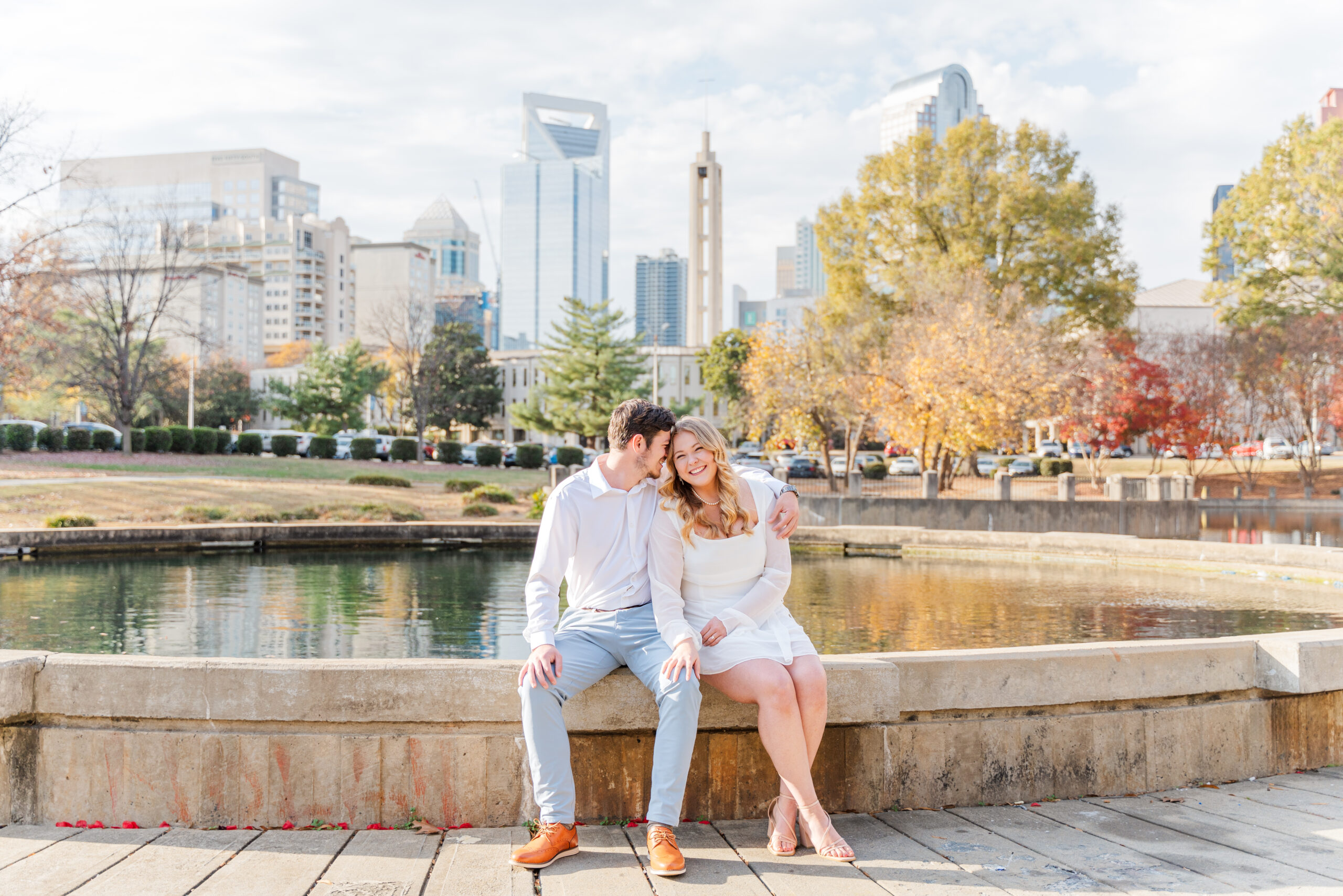 Elaina and Nation sit on the edge of the fountain. The Charlotte, NC city skyline looms behind them and the trees are in full fall color around them.