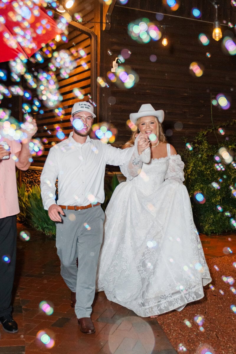 Bride Samantha smiling during bubble fake exit in white cowboy hat and lace gown, Alexander Homestead wedding photos Charlotte NC
