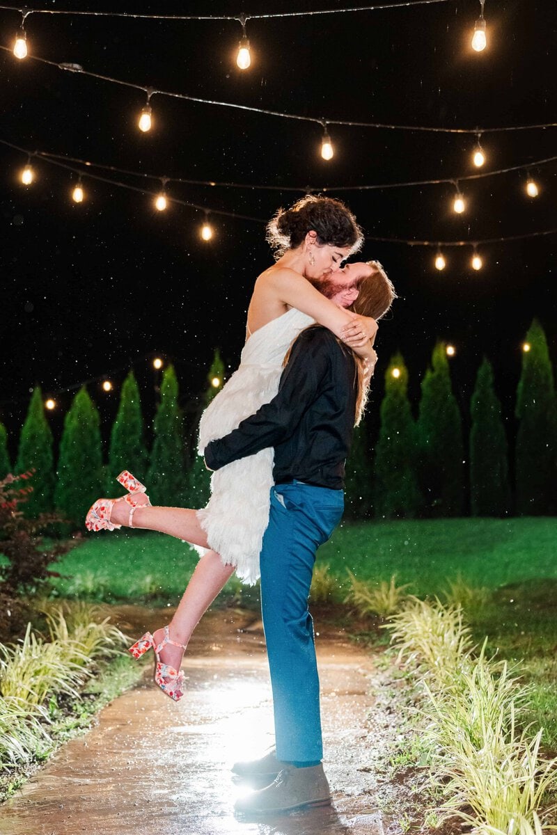Newly weds kiss at night during the rain and under the string lights at mariposa chapel captured by the best charlotte nc wedding photographer