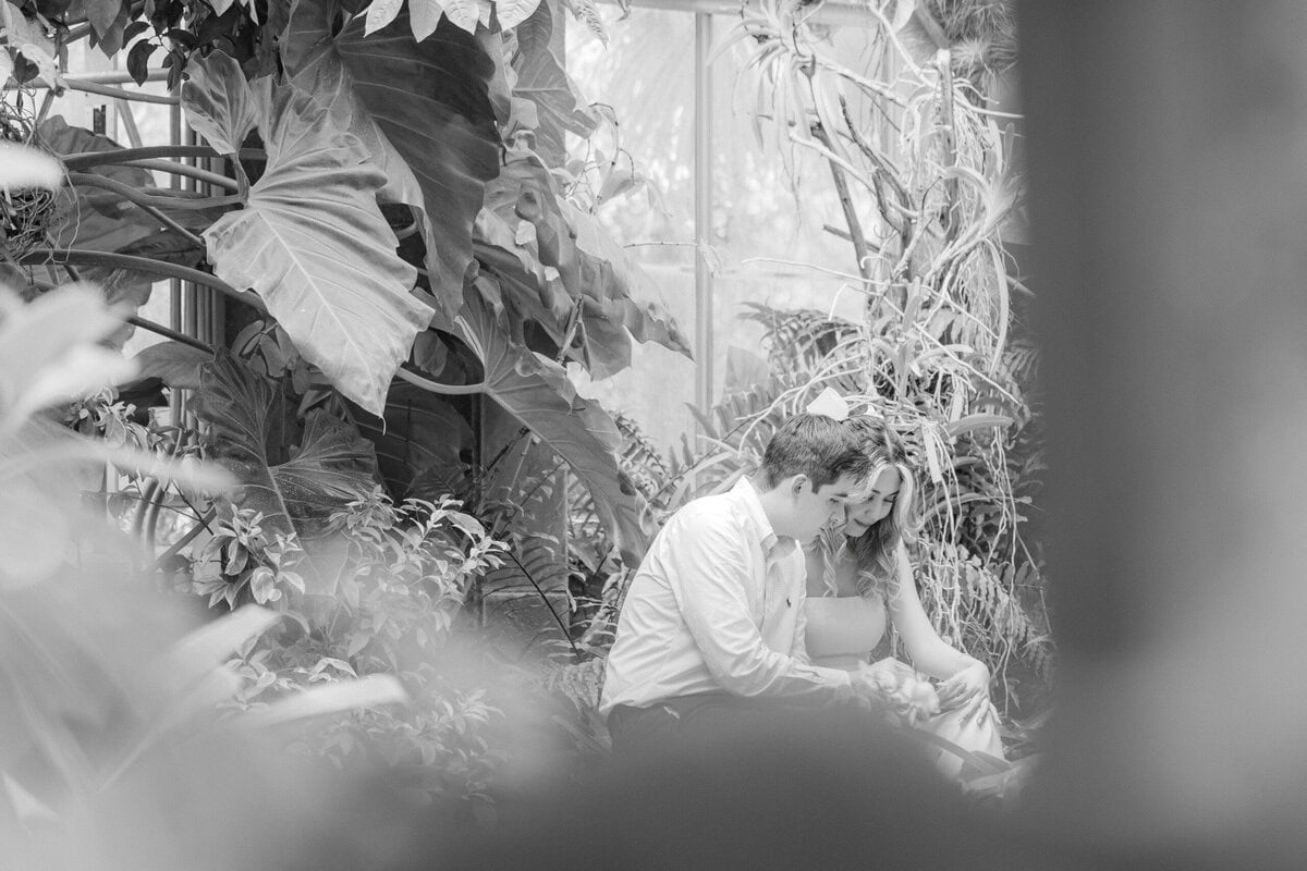 Couple sitting together on a bench inside the Orchid Conservatory at Daniel Stowe Botanical Garden in Belmont NC before a surprise proposal, captured candidly by Charlotte wedding photographer The Lilac Lion Photography