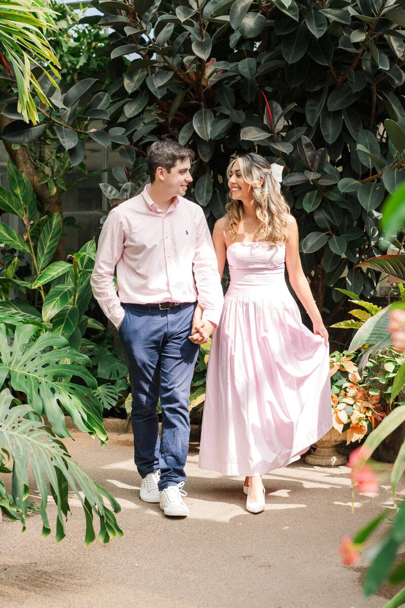 Engaged couple in pale pink outfits posing for portraits inside the Orchid Conservatory at Daniel Stowe Botanical Garden Charlotte area wedding photographer