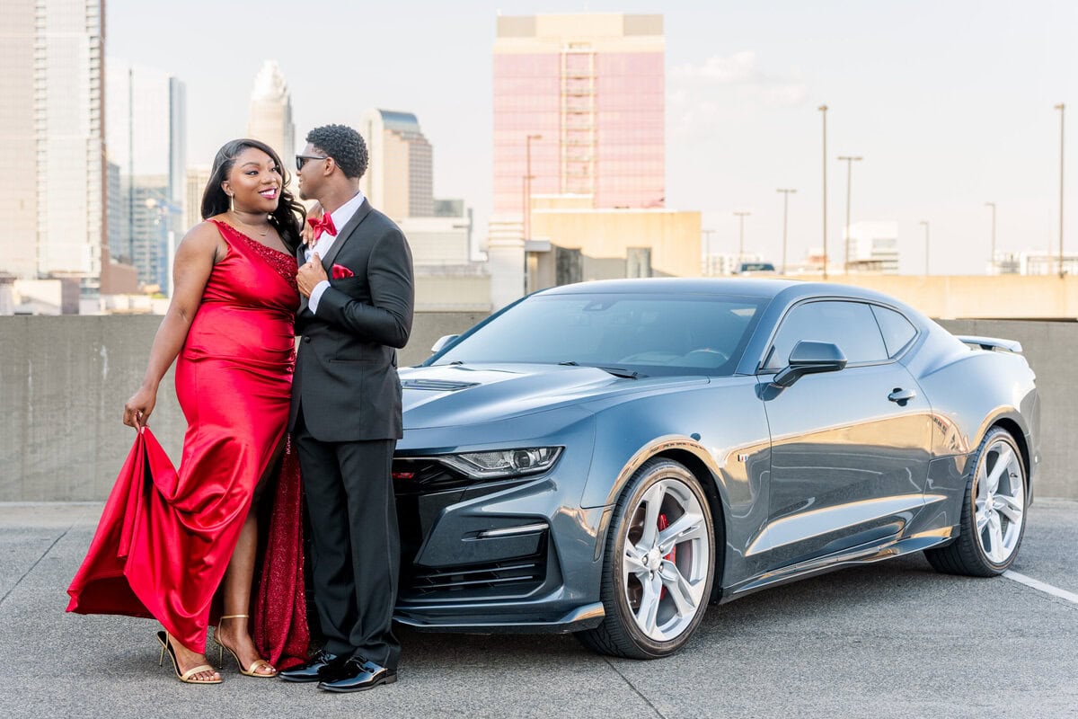 Couple posing with a black sports car on a Southend Charlotte rooftop parking garage with city skyline views