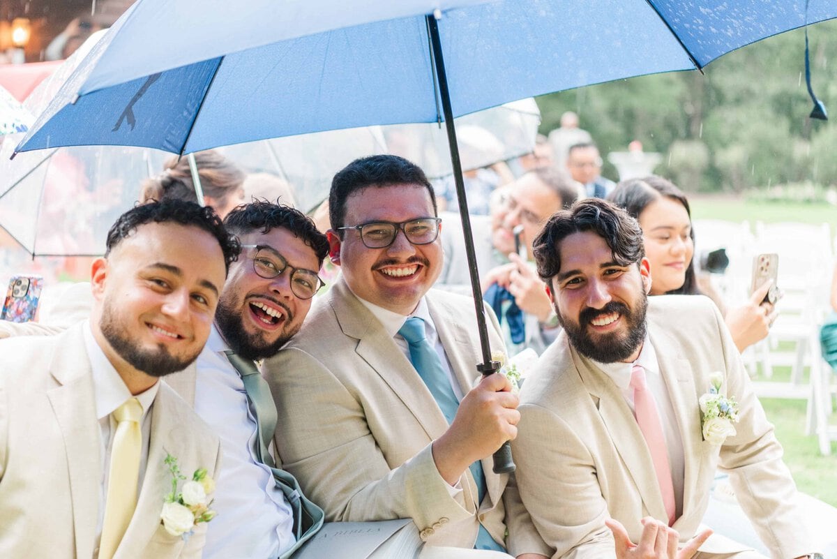 Groomsmen laughing under a blue umbrella in the rain at Rolling Hill Venue