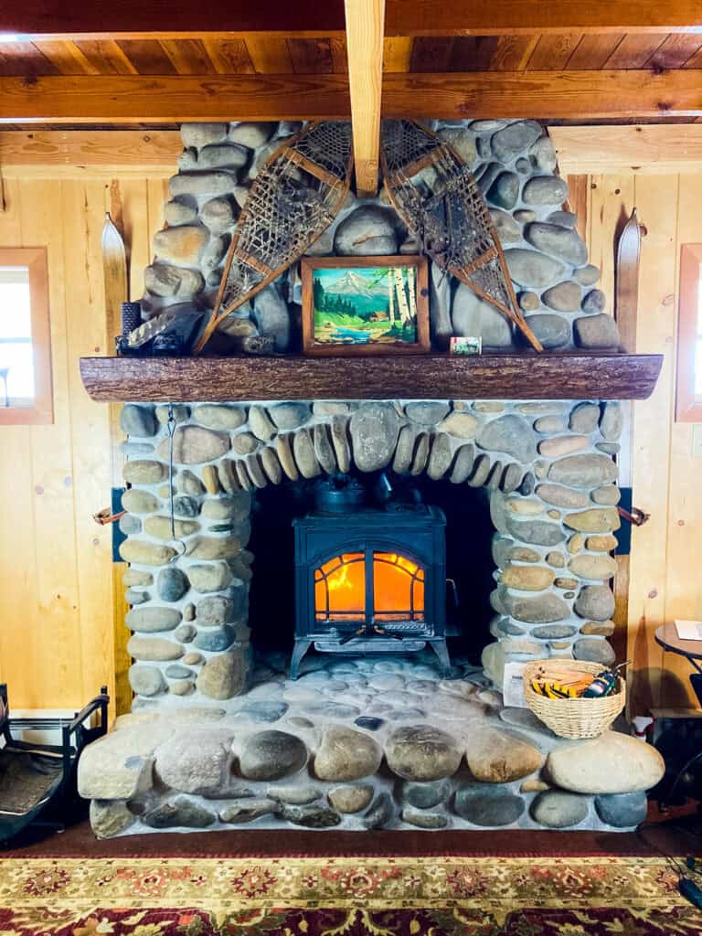 Wood stove in a cabin with pine paneled walls. There is a fire burning. The wood stove is surrounded by a river rock fireplace and wood mantle.