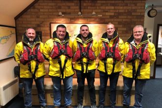 Fishermen in yellow and red safety gear pose indoors, celebrating a rescue award, industry equipment.