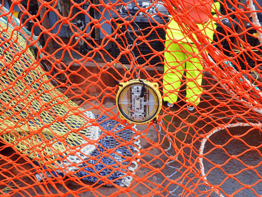 Fishing vessel with gear and crew behind orange netting in commercial fishing industry publication.